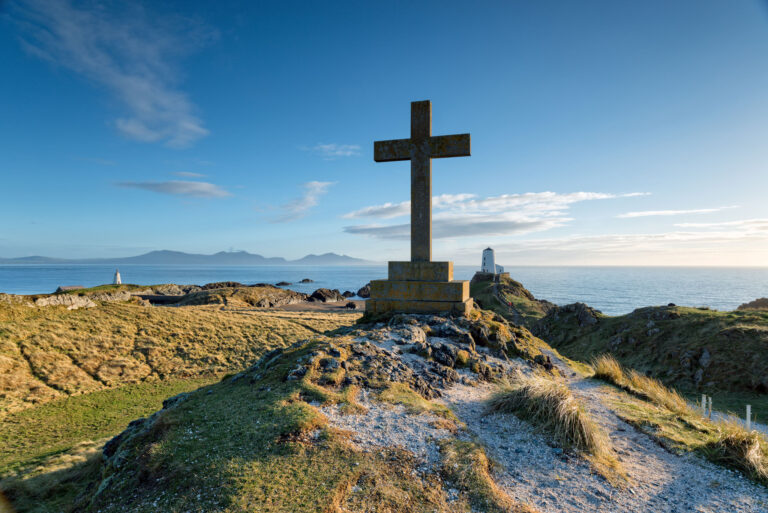 Steinkreuz auf einer Klippe mit Blick auf das Meer bei Sonnenlicht, Symbol für christlichen Glauben und Hoffnung in der Natur.
