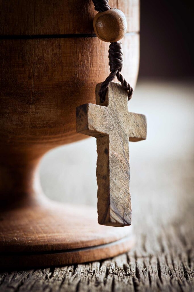 Holzkreuz an einer Kordel vor einem rustikalen Abendmahlskelch – christliches Symbol für Glaube, Erlösung und das Abendmahl.