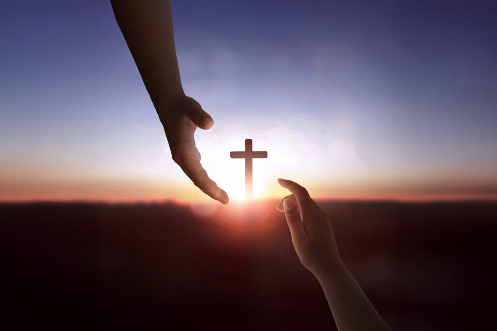 Zwei Hände greifen symbolisch nach einem leuchtenden Kreuz im Sonnenuntergang – christliches Motiv für Glaube, Hoffnung und Verbindung zu Gott.