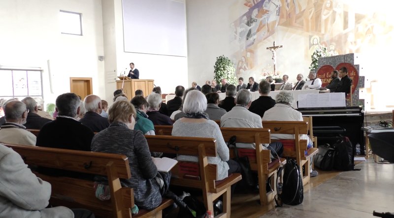 Teilnehmer der Kirchweihmontagskonferenz sitzen in Holzbankreihen in einer hellen Kirche und hören einem Redner an der Kanzel zu, hinter dem ein Kirchenaltar mit Kreuz und Wandgemälde zu sehen ist.