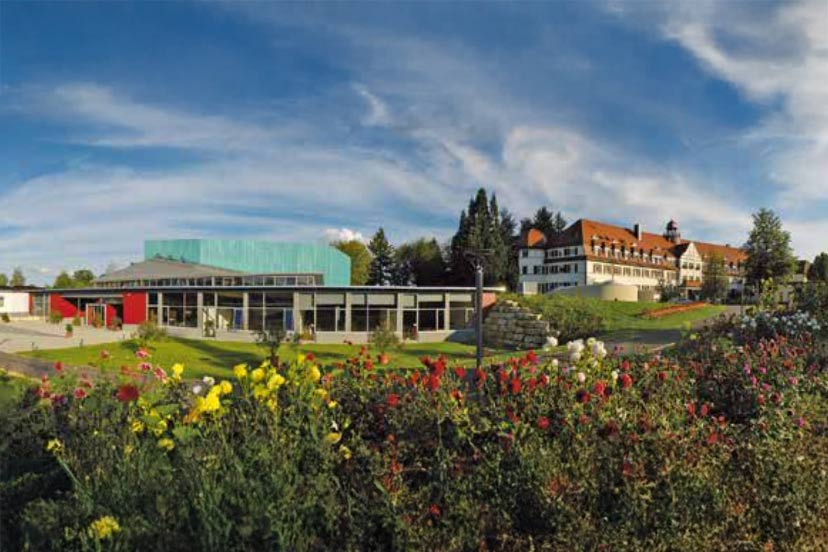 Panorama des Forum Schönblick Tagungshauses in Schwäbisch Gmünd: im Vordergrund bunte Blumenrabatten, im Hintergrund ein modernes Konferenzgebäude neben einem historischen Bildungs- und Gästehaus unter blauem Himmel.