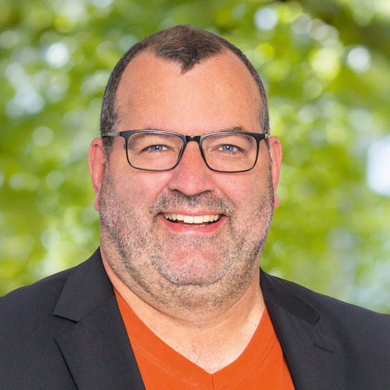 Porträt von Matthias Hanssmann, Mitglied der Leitung der ChristusBewegung Lebendige Gemeinde – freundlich lächelnder Mann mit Brille und Bart vor grünem, natürlichem Hintergrund.