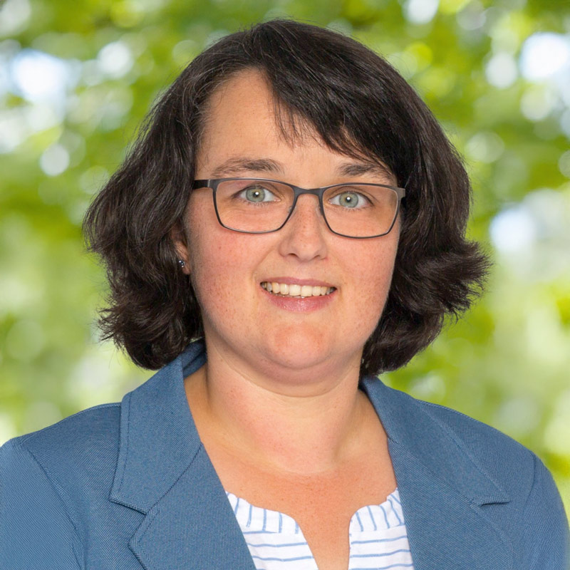 Porträt von Anja Holland, Mitglied der Leitung der ChristusBewegung Lebendige Gemeinde – Frau mit dunklem Haar und Brille, freundlich lächelnd vor grünem, natürlichem Hintergrund.