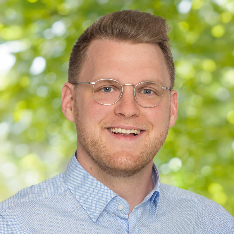 Porträt von Christian Nathan, Mitglied der Leitung der ChristusBewegung Lebendige Gemeinde – junger Mann mit Brille und Bart, freundlich lächelnd vor grünem Hintergrund.