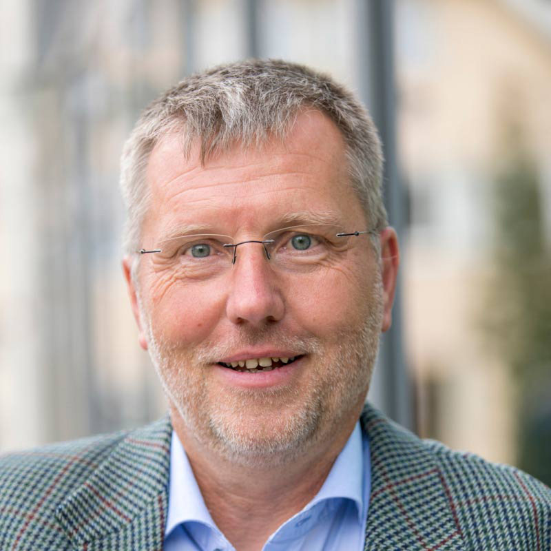 Porträt von Dieter Schenk, Vorstandsmitglied der ChristusBewegung Lebendige Gemeinde, mit kariertem Sakko, blauem Hemd und Brille vor verschwommenem Hintergrund im Freien.